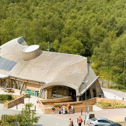 Modern visitor centre with curved wooden roof and solar panels, surrounded by trees, with people and cars in the foreground.