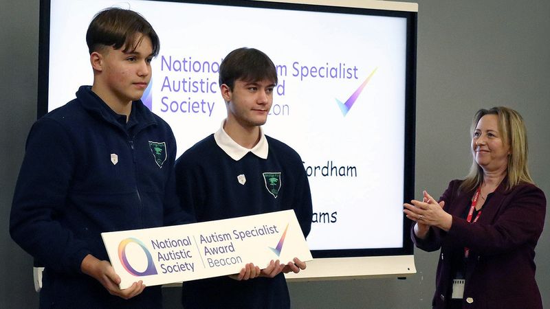 Two teenage boys in Grange Park uniform holding a plaque . Beverley Fordham is stood on the right clapping after presenting them with the award