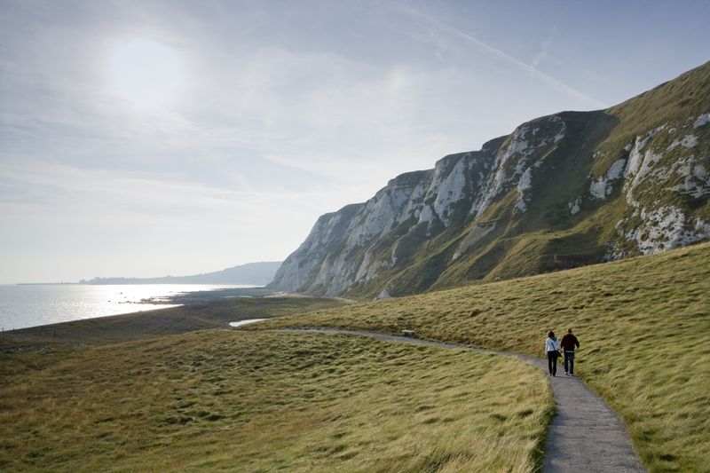 Two people walking along a path beside white cliffs with a sunlit sea in the distance
