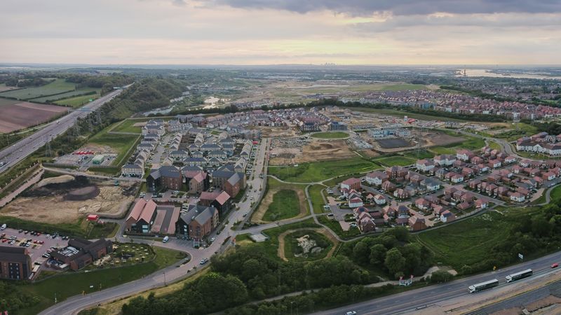 Aerial view of Ebbsfleet Garden City showing a mix of newly built homes, residential streets, green spaces and ongoing construction areas. Roads and a large roundabout run through the development, with surrounding countryside and the River Thames visible in the distance beneath a cloudy sky.