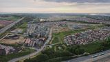 Aerial view of Ebbsfleet Garden City showing a mix of newly built homes, residential streets, green spaces and ongoing construction areas. Roads and a large roundabout run through the development, with surrounding countryside and the River Thames visible in the distance beneath a cloudy sky.
