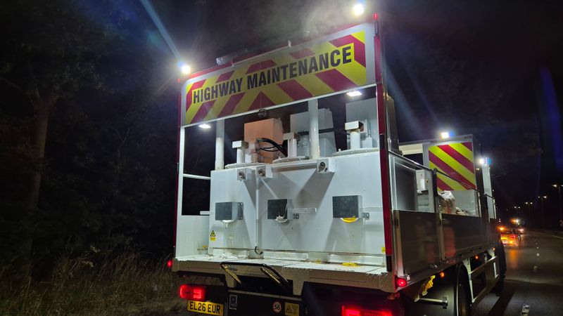 "A highway maintenance vehicle positioned on a road during overnight maintenance works, with reflective safety markings and illuminated warning signs visible."