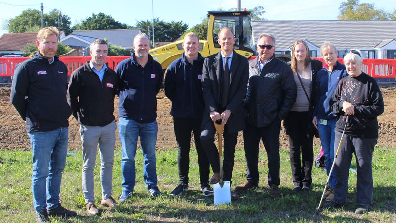 Groundbreaking ceremony. 9 people standing in a line, the middle holding a shovel with construction machinery in the background.