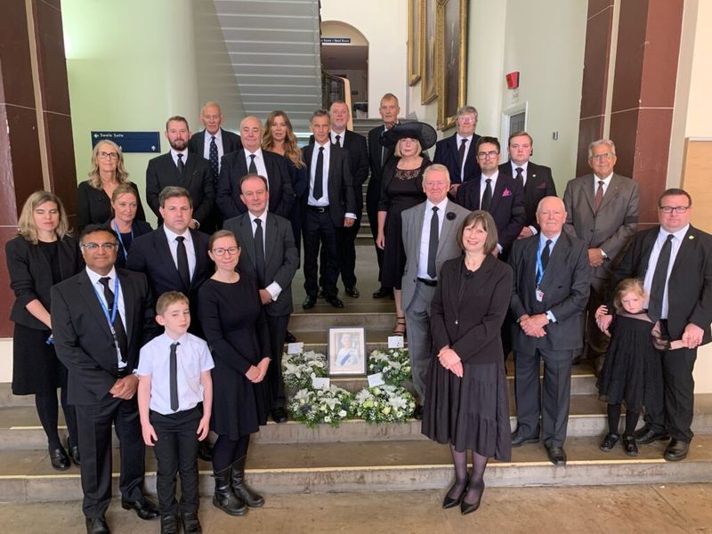 Image of Directors and Members all in funeral clothing, standing on the County Hall step surrounding four wreaths, which circle a picture of the late Queen Elizabeth the Second