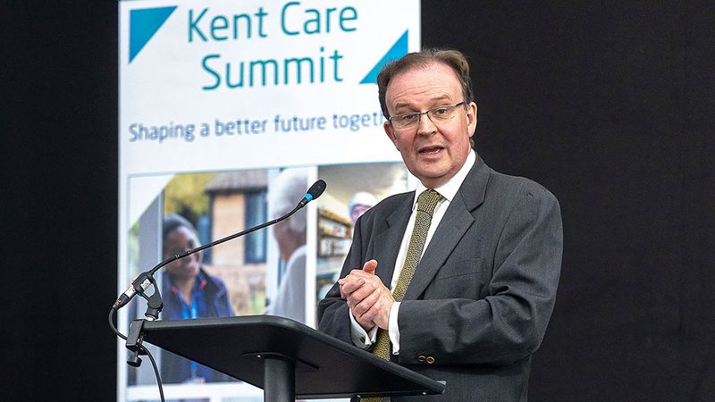 Roger Gough, KCC Leader, at lectern on stage with the words 'Kent Care Summit - shaping a better future together'  on a screen behind him.
