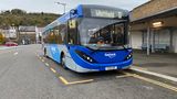 A photograph of a blue bus on a grey road. People are waiting to get on the bus. The bus says 'Whitfield' on it.