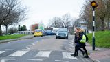 Two school children cross the road at a zebra crossing. One of them is holding a bright, reflective backpack.