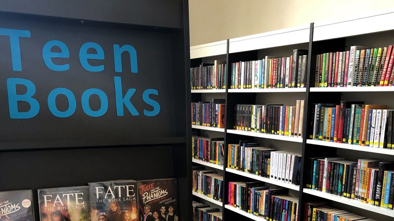 A book shelf with the words 'Teen Books' in blue on the top with a range of books underneath. More bookshelves with books organised by category in the background 