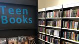A book shelf with the words 'Teen Books' in blue on the top with a range of books underneath. More bookshelves with books organised by category in the background 