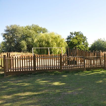 Wooden fenced play area, surrounded by trees and greenery under a clear blue sky.