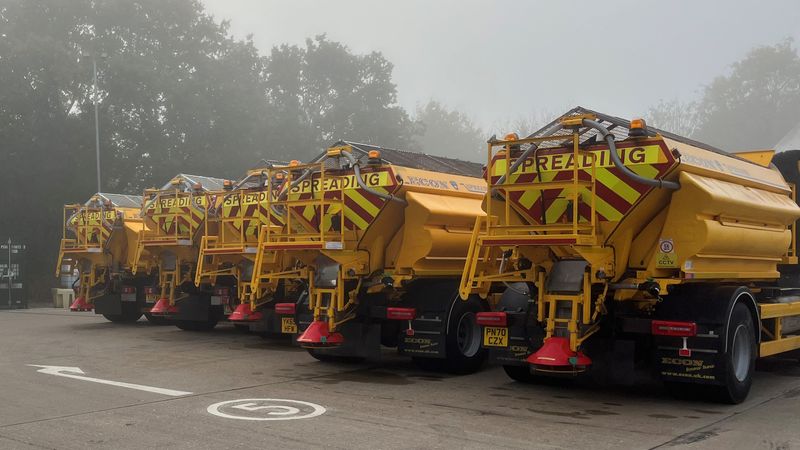 A row of bright yellow gritting trucks parked in formation on a foggy morning at a depot.