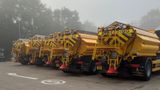 A row of bright yellow gritting trucks parked in formation on a foggy morning at a depot.