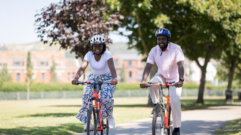 Two people wearing helmets cycle side by side on a paved path through a green park, with trees and buildings visible in the background.