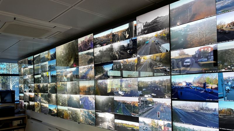Large wall of multiple CCTV screens showing live views of roads, intersections, and traffic from different locations inside a monitoring control room.