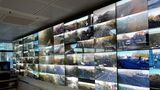 Large wall of multiple CCTV screens showing live views of roads, intersections, and traffic from different locations inside a monitoring control room.