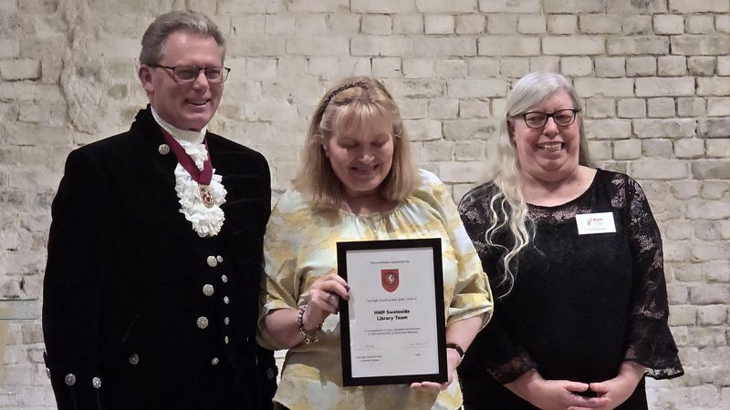 Three people stand in front of a light brick wall at an award event. The central person holds a framed certificate reading &ldquo;HMP Swaleside Library Team,&rdquo; presented by the High Sheriff of Kent. One person on the left wears a formal black outfit with a ceremonial chain, while the person on the right wears a black lace top with a name badge.