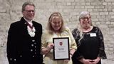 Three people stand in front of a light brick wall at an award event. The central person holds a framed certificate reading &ldquo;HMP Swaleside Library Team,&rdquo; presented by the High Sheriff of Kent. One person on the left wears a formal black outfit with a ceremonial chain, while the person on the right wears a black lace top with a name badge.