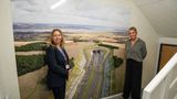 Two people standing indoors in front of a large wall graphic showing an aerial view of the Lower Thames Crossing road tunnel and surrounding landscape.