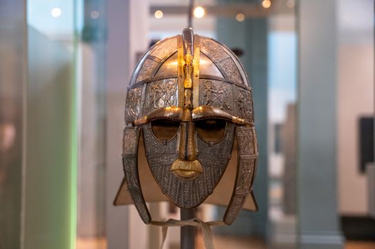 Close-up of an ornate Anglo-Saxon helmet on display, featuring a metal face mask with detailed engravings and decorative panels.