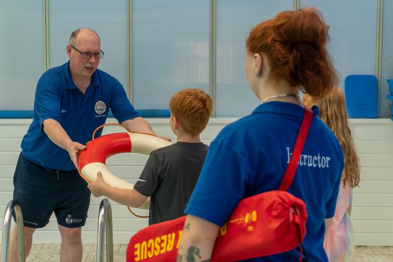 A life-saving instructor at an indoor swimming pool hands a red and white lifebuoy to a young boy. Two other young people look on. 