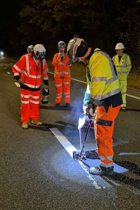 "Road workers in high-visibility clothing applying fresh white road markings on a newly resurfaced road during overnight maintenance work."