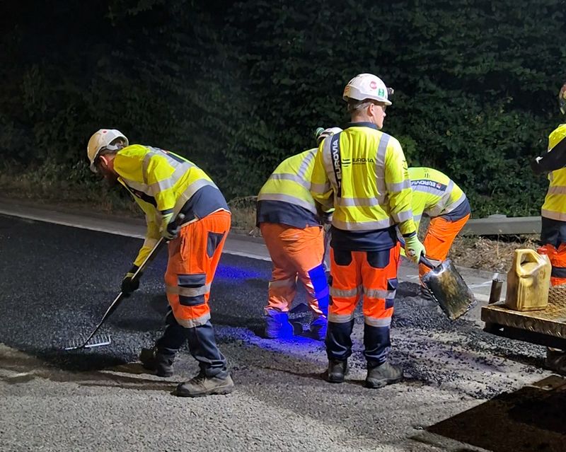 Road workers in high-visibility jackets and hard hats resurface a road at night, spreading and levelling fresh asphalt under artificial lighting. A tree-lined embankment forms the background, while equipment and materials are visible at the roadside.