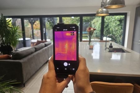 A thermal imaging camera attached to a smartphone, held in a living room with large glass doors overlooking a garden.
