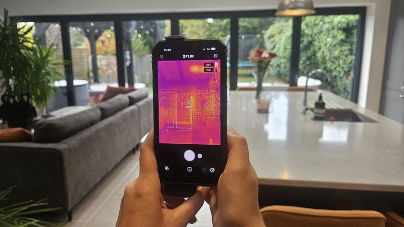 A thermal imaging camera attached to a smartphone, held in a living room with large glass doors overlooking a garden.